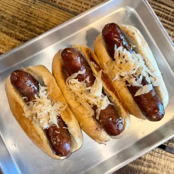 Three smoked bratwursts on toasted buns topped with sauerkraut, lined up on a metal tray