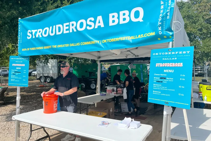 The Strouderosa BBQ tent at Oktoberfest Dallas on Flag Pole Hill, a teal Strouderosa BBQ banner overhead, menu boards on the tent poles and the crew working behind the front table