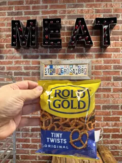 A bag of Rold Gold Tiny Twists pretzels held up in front of the MEAT marquee sign and the Strouderosa BBQ license plate board